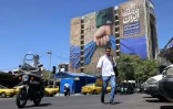 Des passants traversent une rue devant un panneau d'affichage apposé sur la façade d'un immeuble représentant le détroit d'Ormuz, avec une légende en persan indiquant "Pour toujours entre les mains de l'Iran", à Téhéran, le 25 mai 2026