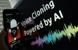 Photo d'illustration montrant un téléphone sur lequel une application dictaphone est ouverte, avec un écran à l'arrière-plan sur lequel on peut lire "Voice Cloning Powered by AI"
