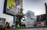 Un policier décharge d'un camion des urnes destinés à un bureau de vote devant une affiche électorale du président sortant Yoweri Museveni, candidat du Mouvement de résistance nationale (NRM) à la présidentielle, le 15 janvier 2026 à Kampala, en Ouganda