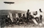 Reproduction d'une carte postale, issue de la collection de l'Historial de Péronne, montrant des Poilus tirant au canon tandis qu'un zeppelin, dirigeable allemand, survole le champ de bataille