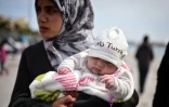 Dans les bras de sa mère migrante, un bébé porte un bonnet où il est écrit "No Turkey"