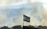 Un drapeau israélien flotte devant des panaches de fumée qui s'élèvent d'un champ après que des roquettes lancées depuis le Sud-Liban ont atterri près de Katzrin, sur le plateau du Golan annexé par Israël, le 13 juin 2024