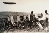 Reproduction d'une carte postale, issue de la collection de l'Historial de Péronne, montrant des Poilus tirant au canon tandis qu'un zeppelin, dirigeable allemand, survole le champ de bataille