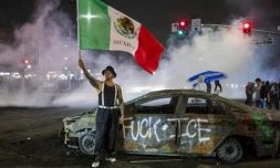 Un manifestant tient un drapeau mexicain devant l'épave d'une voiture incendiée lors de heurts avec les forces de l'ordre contre l'expulsion d'immigrés, à Los Angeles, le 7 juin 2025 en Californie
