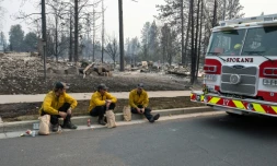 Trois membres du service d'incendie de Spokane en pause sur le bord du trottoir devant des maisons d'habitation incendiées, dans le quartier de Balboa à Spokane (Washington), le 2 août 2026