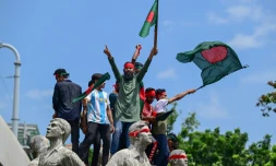 Des manifestants juchés sur un monument à la mémoire des victimes du terrorisme brandissent des drapeaux bangladais, le 4 août 2024 à Dacca