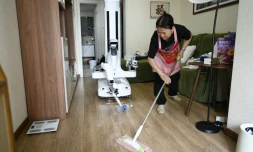 Un robot assistant ménager transporte une bouteille jusqu'à une poubelle tandis qu'une femme de ménage nettoie le sol chez une cliente à Pékin, le 21 mai 2026 en Chine