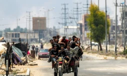 Des hommes à l'arrière d'un tricycle le long de la route de Salaheddin à Deir al-Balah, dans le centre de la bande de Gaza, le 21 juillet 2025, tandis que derrière eux s'élèvent des volutes de fumée provenant des bombardements israéliens