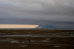 Un homme marche sur une plage d'Andernos au bord du bassin d'Arcachon, au fond duquel s'élèvent les fumées de l'incendie de Biscarosse, le 23 juillet 2026