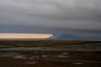 Un homme marche sur une plage d'Andernos au bord du bassin d'Arcachon, au fond duquel s'élèvent les fumées de l'incendie de Biscarosse, le 23 juillet 2026