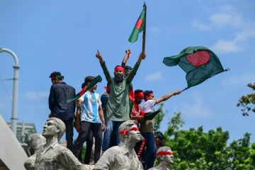 Des manifestants juchés sur un monument à la mémoire des victimes du terrorisme brandissent des drapeaux bangladais, le 4 août 2024 à Dacca