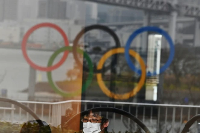 Les anneaux olympiques se réflétant sur une vitrine dans le parc de bord de mer d'Odaiba (baie de Tokyo) et un Japonais se protégeant du nouveau coronavirus avec un masque, le 8 mars 2020