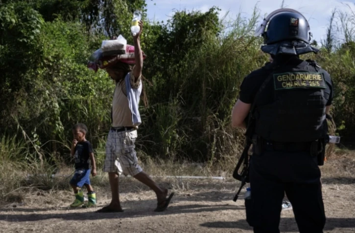 Un habitant, transportant sur sa tête des sacs de provisions, passe avec des enfants, devant un gendarme à un barrage routier à Saint-Louis, au sud de Nouméa, le 23 septembre 2024 en Nouvelle-Calédonie ( AFP / SEBASTIEN BOZON )