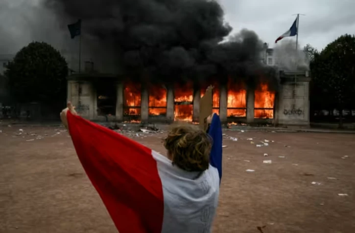 Un protestataire enveloppé dans un drapeau français devant l'antenne de l'Office du tourisme de Lyon en feu, lors d'une manifestation contre la vie chère, le 17 octobre 2026 ( AFP / Jeff PACHOUD )