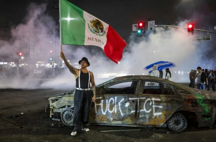 Un manifestant tient un drapeau mexicain devant l'épave d'une voiture incendiée lors de heurts avec les forces de l'ordre contre l'expulsion d'immigrés, à Los Angeles, le 7 juin 2025 en Californie