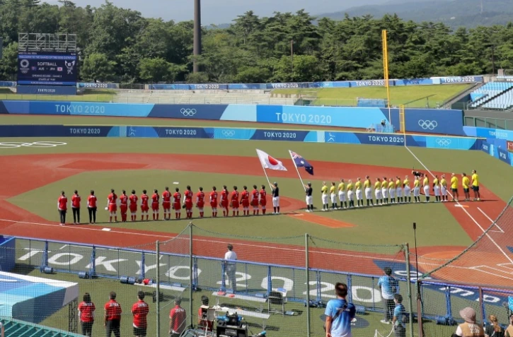 Les équipes japonaises et australiennes de softball écoutent les hymnes nationaux avant le début du premier match de la compétition olympique, marquant le début officiel des épreuves des JO de Tokyo, le 21 juillet 2021 à Fukushima, à deux jours de la cérémonie d'ouverture