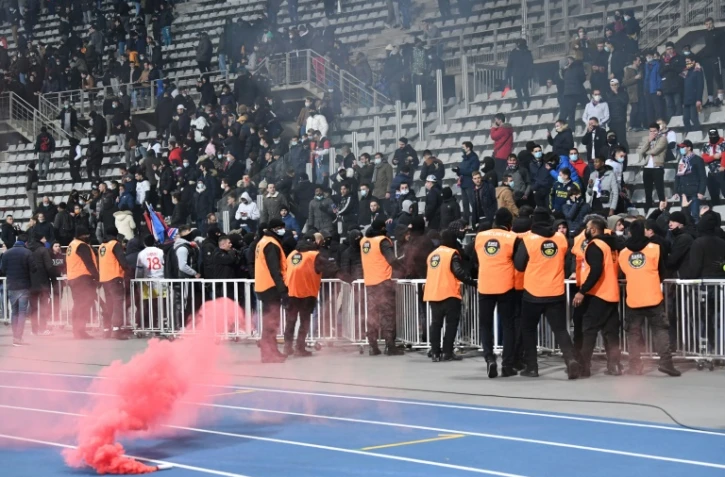Un fumigène jeté sur la piste du stade Charléty tandis que des stadiers font face aux supporters, lors du 32e de finale de Coupe de France entre le Paris FC et Lyon le 17 décembre 2021, définitivement interrompu à la mi-temps en raison des incidents en tribunes