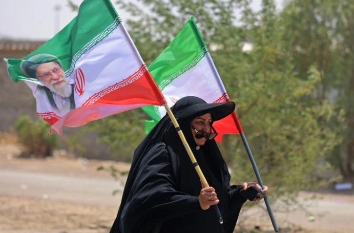 Une pèlerine iranienne brandit des drapeaux iraniens dont l'un comprend un portrait du guide suprême Ali Khamenei, tué dans un bombardement américain, le 29 juillet 2026 dans la ville-frontière de Zurbatiyah, en Irak