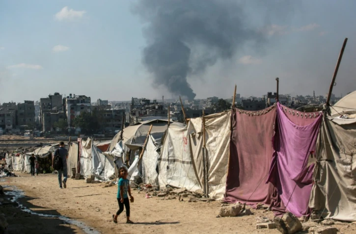 Des Palestiniens devant des tentes à Gaza-ville avec en arrière-plan une colonne de fumée s'élevant du centre de la bande de Gaza après un bombardement israélien, le 6 juillet 2025