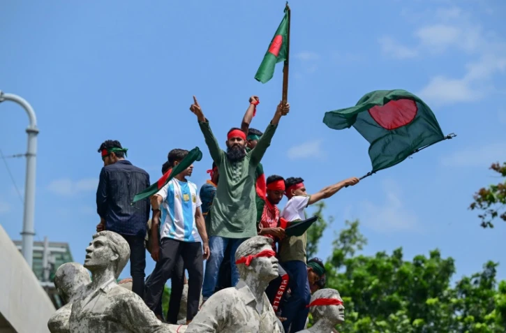 Des manifestants juchés sur un monument à la mémoire des victimes du terrorisme brandissent des drapeaux bangladais, le 4 août 2024 à Dacca