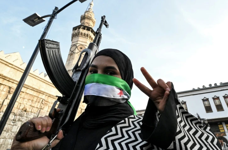 Une femme masquant son visage avec le drapeau de l'opposition syrienne fait le V de la victoire en tenant un fusil d'assaut devant la mosquée des Omeyyades à Damas le 10 décembre 2024