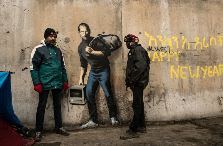 Des migrants devant une oeuvre de l'artiste britannique Bansky photographiée le 12 décembre 2015 et située à l'entrée de la "Jungle" de Calais, représente Steve Jobs (Apple), qui porte un baluchon et un vieil ordinateur