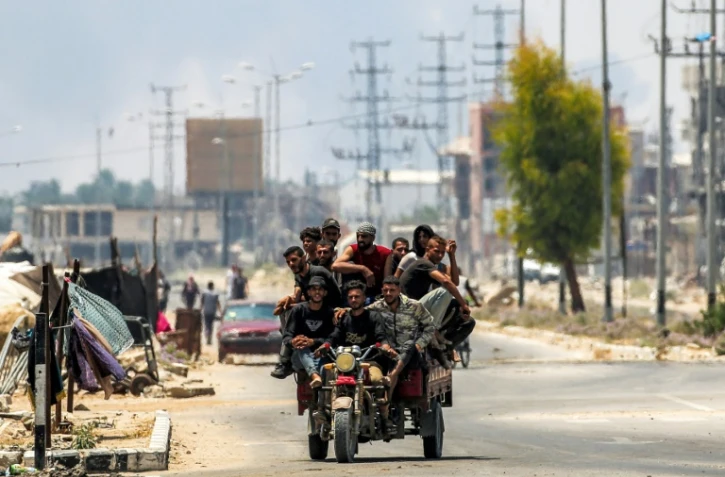 Des hommes à l'arrière d'un tricycle le long de la route de Salaheddin à Deir al-Balah, dans le centre de la bande de Gaza, le 21 juillet 2025, tandis que derrière eux s'élèvent des volutes de fumée provenant des bombardements israéliens
