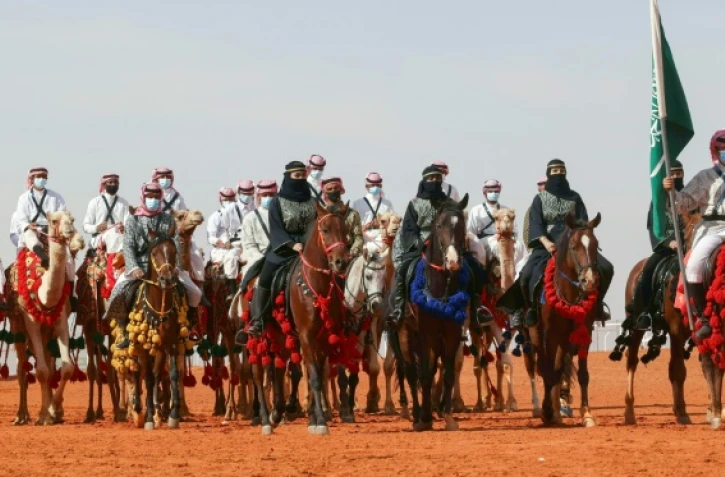 Des femmes en niqab noir devancent à cheval des hommes en robe blanche à dos de chameaux lors de festivités dans le cadre d'un concours de beauté pour camélidés réservé aux femmes dans le désert de Rumah, le 8 janvier 2022