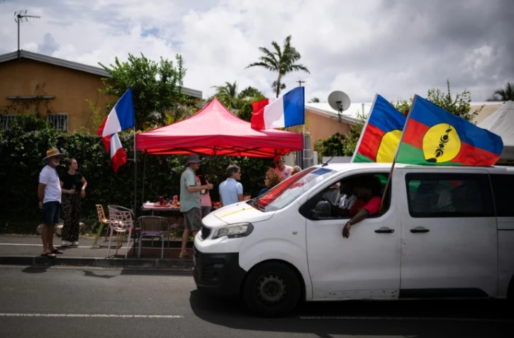 Des habitants célèbrent l'anniversaire de la prise de possession de la Nouvelle-Calédonie par la France, tandis qu'une voiture occupée par des indépendantiste passe dans une rue de Nouméa, dans le quartier de Magenta, le 24 septembre 2024