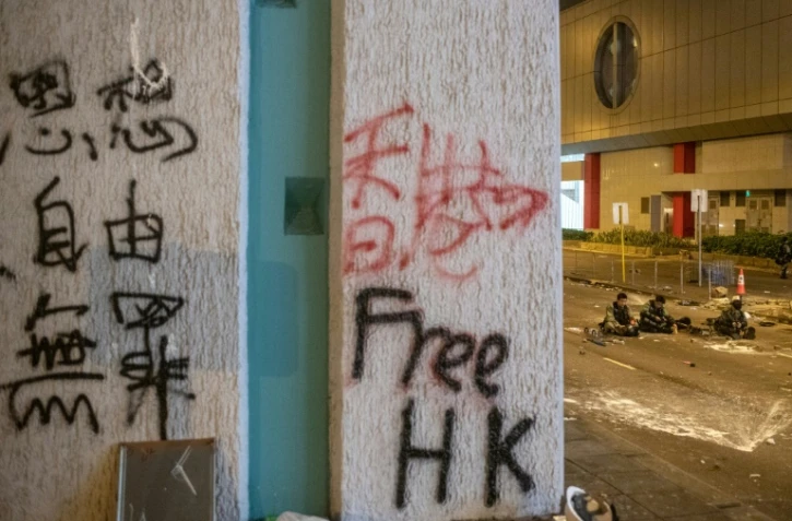 Un graffiti sur un mur après des affrontements entre des manifestants et des policiers anti-émeutes qui se reposent à l'arrière-plan (D), à Hong Kong au petit matin du 22 juillet 2019