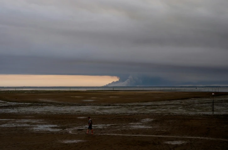 Un homme marche sur une plage d'Andernos au bord du bassin d'Arcachon, au fond duquel s'élèvent les fumées de l'incendie de Biscarosse, le 23 juillet 2026