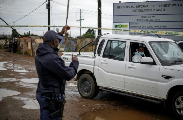 Un policier sud-africain soulève la bande jaune barrant l'accès pour permettre à un expert de la police scientifique d'approcher la scène d'une tuerie dans un bar clandestin, le 21 décembre 2025 à Bekkersdal (Afrique du Sud)