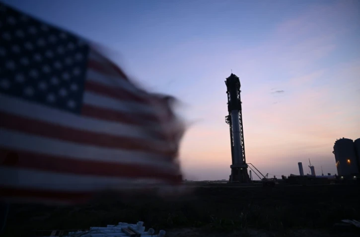 Un drapeau amércain flottant au vent, avec en arrière plan la fusée de SpaceX Starship debout sur son pas de tir à Boca Chica au Texas, le 18 avril 2023
